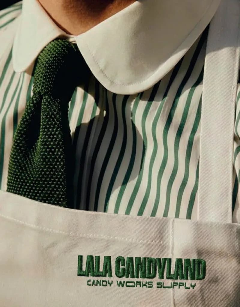 A white LaLa Candyland apron, embroidered in green with the logo and “Candy Works Supply”, worn over a green-and-white striped shirt with a rounded collar and a knitted green tie.